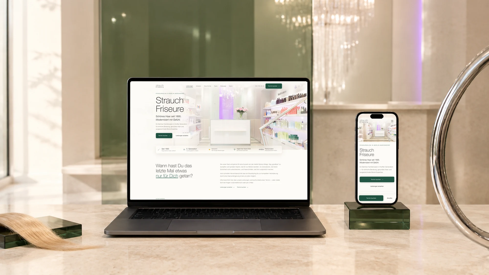 Der neue STRAUCH-Auftritt im Salon aufgenommen: auf dem Laptop die Startseite mit dem Empfangsbereich, der Überschrift „Strauch Friseure“ und der Schaltfläche „Termin buchen“, daneben dieselbe Startseite auf einem Smartphone